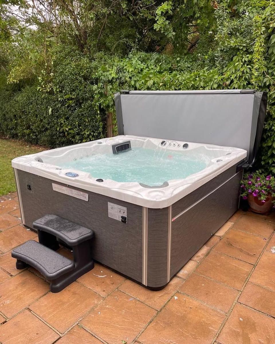 Hot tub on a patio, gray exterior, cover open, steps in front. Green foliage background.