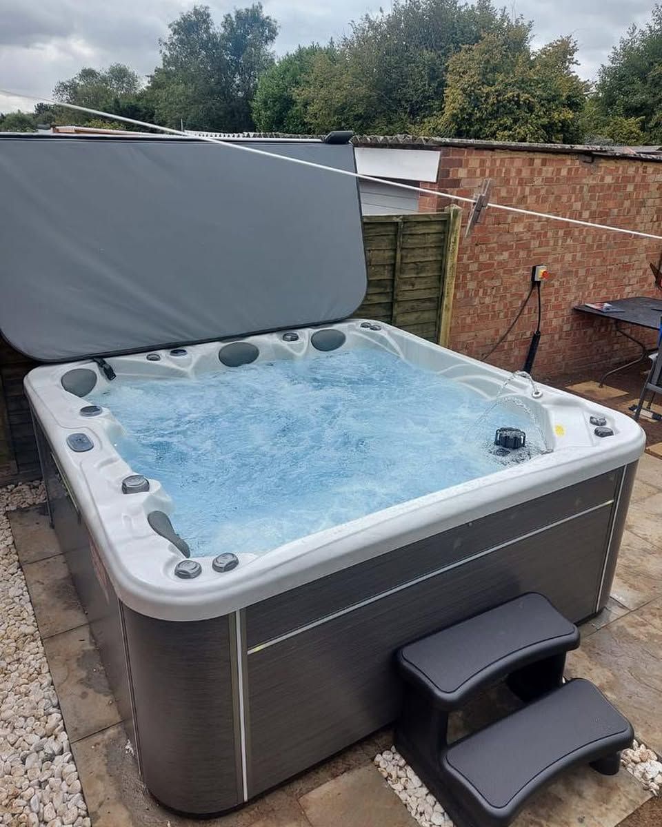 Hot tub with water and cover open; steps on the side. Outdoors setting, gray and tan colors.