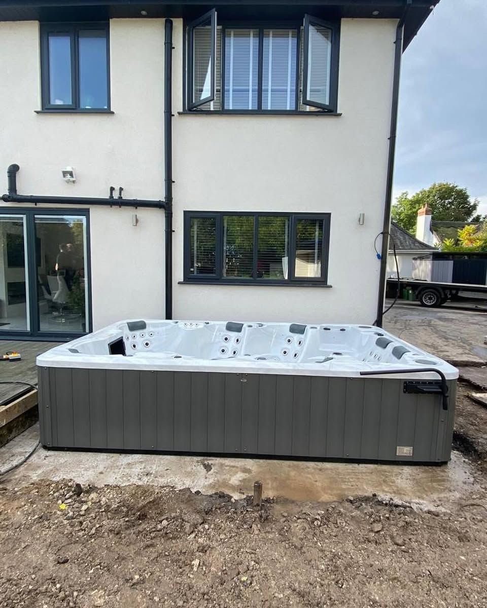 Hot tub against the back of a white house with dark framed windows. The tub is gray, on a concrete base.