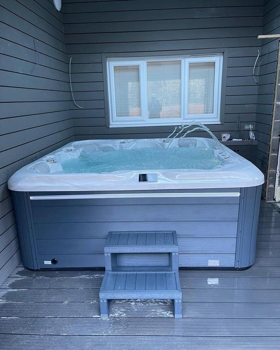 Hot tub with step stool on a deck, under a window. Gray siding and steps. Water is bubbling.