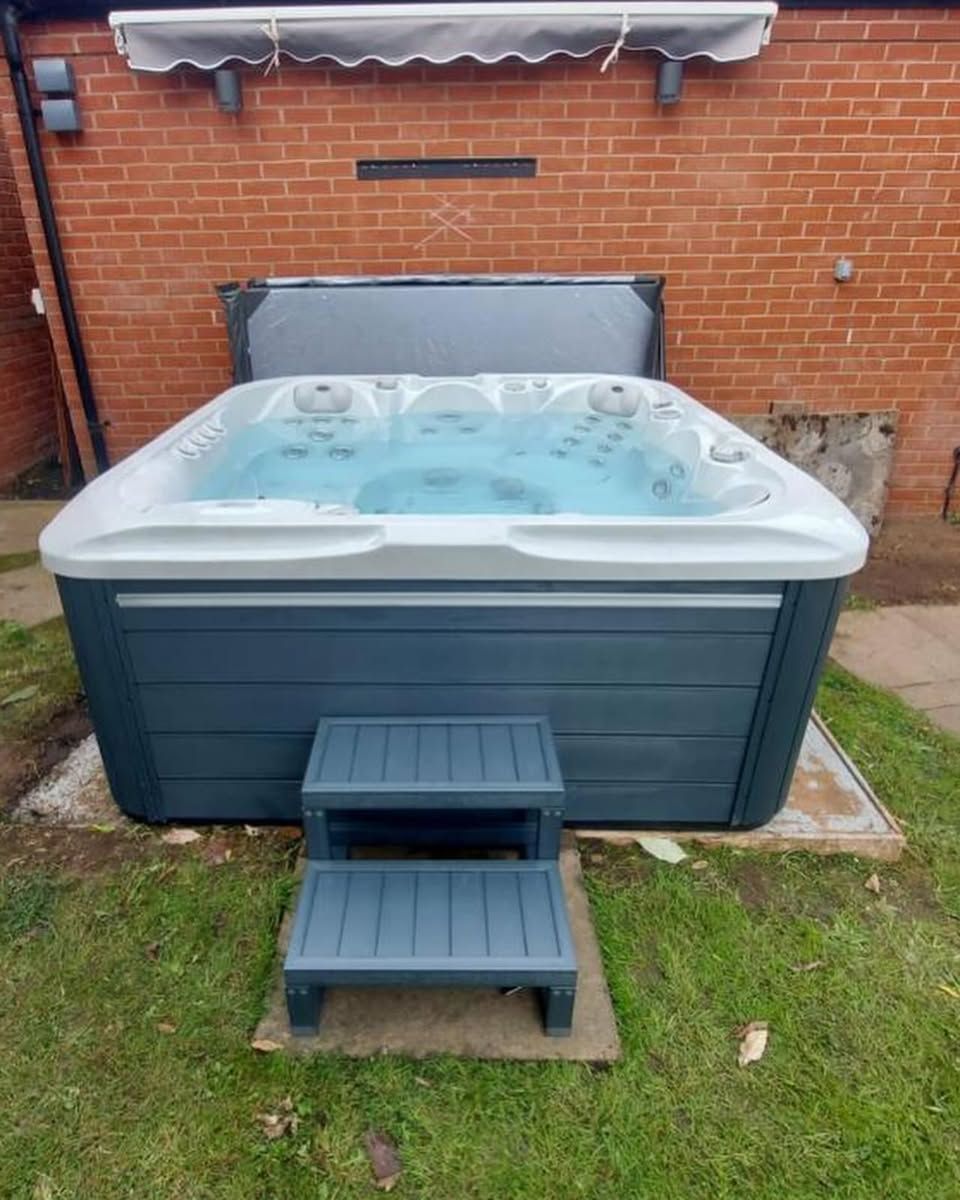 A blue hot tub with steps on a concrete pad, backed by a brick wall and awning.