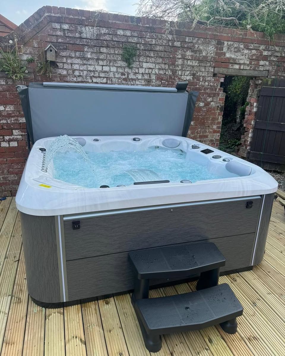 Hot tub with partially open cover, on wooden deck, next to a brick wall. Black step stool in front.