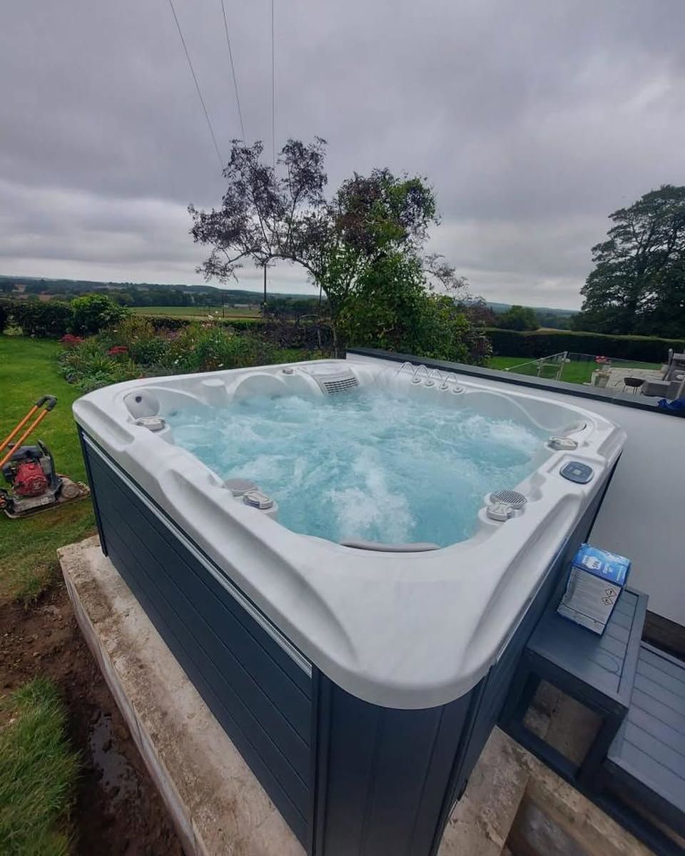 Hot tub with bubbling water on a concrete base, set against a green landscape under a cloudy sky.