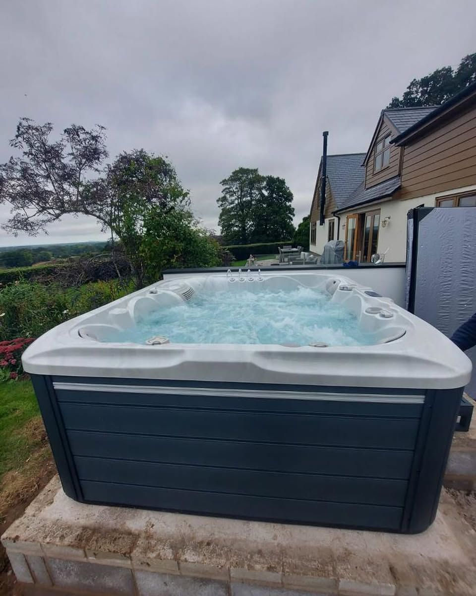 Hot tub with bubbling water sits on a stone base, near a house and trees, under a cloudy sky.