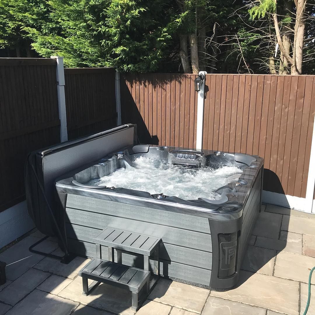 Hot tub on stone patio, cover open. Dark gray spa with bubbling water, small steps, and brown fence background.