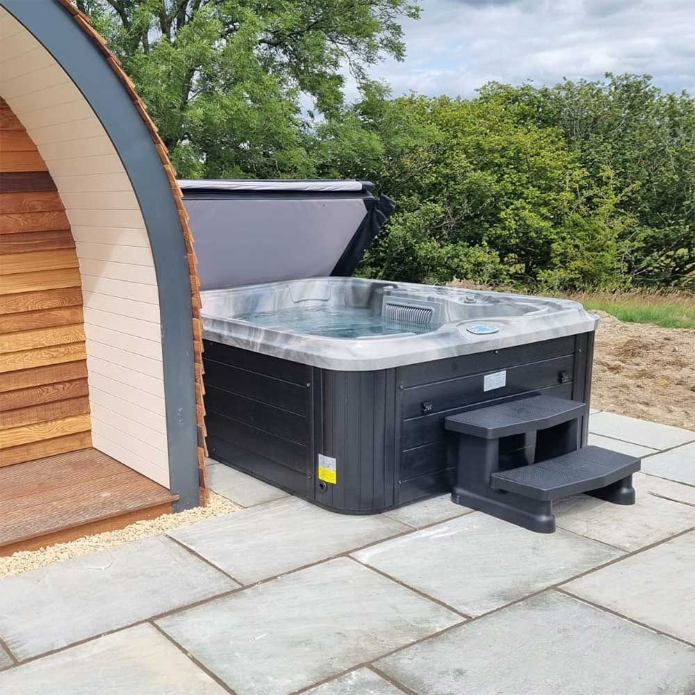 Hot tub with open cover next to a wooden pod, set on stone pavers, with green bushes in the background.