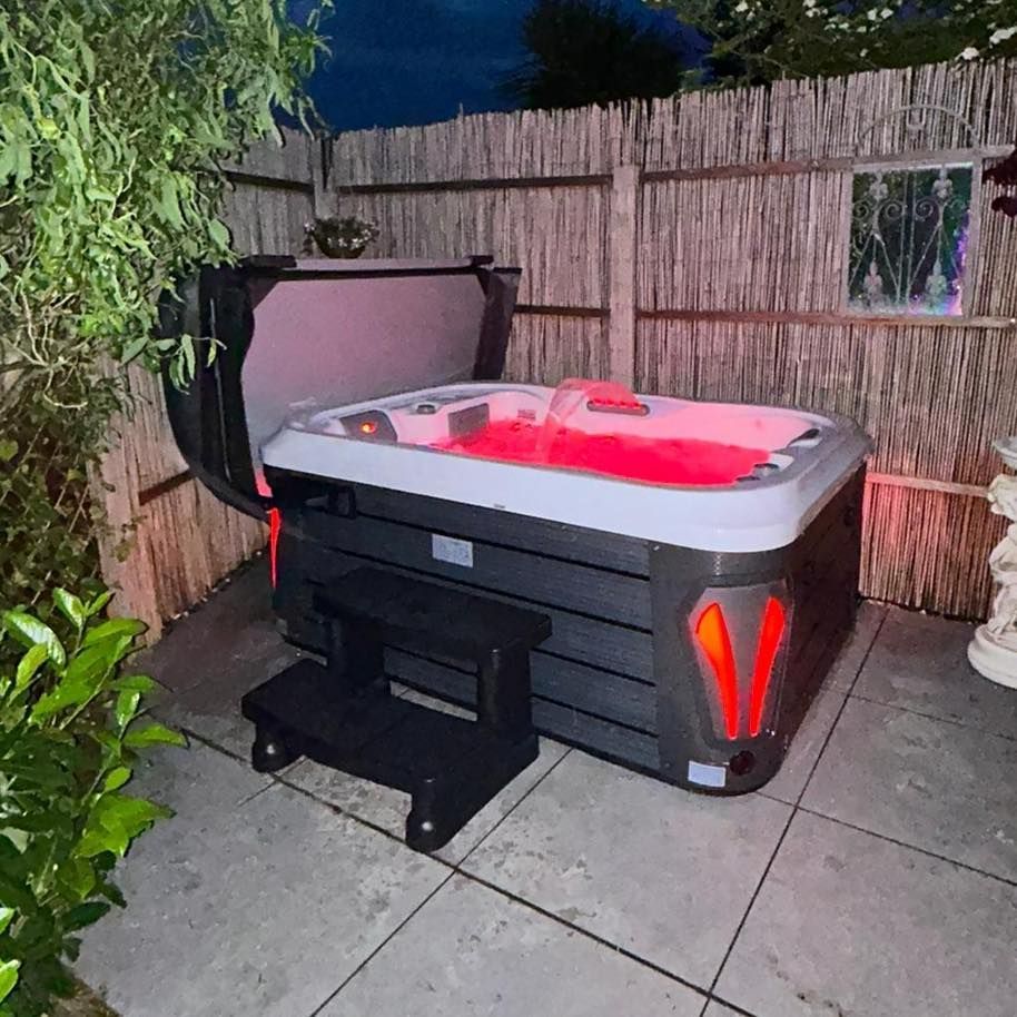 Hot tub with red lights and opened cover, beside a bamboo fence, at dusk. Black steps are in front.
