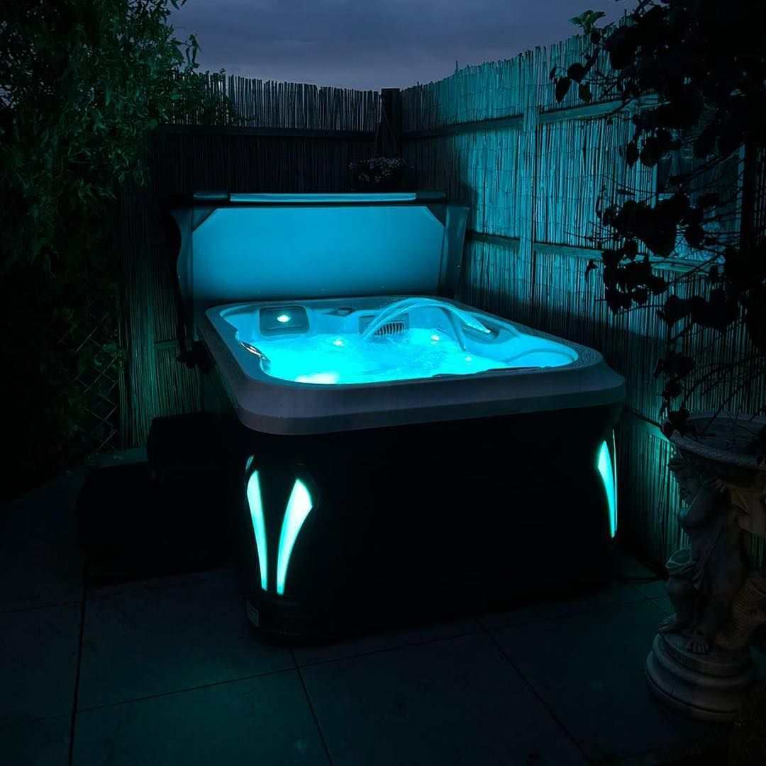 Lit-up outdoor hot tub at night, surrounded by plants and bamboo fence.