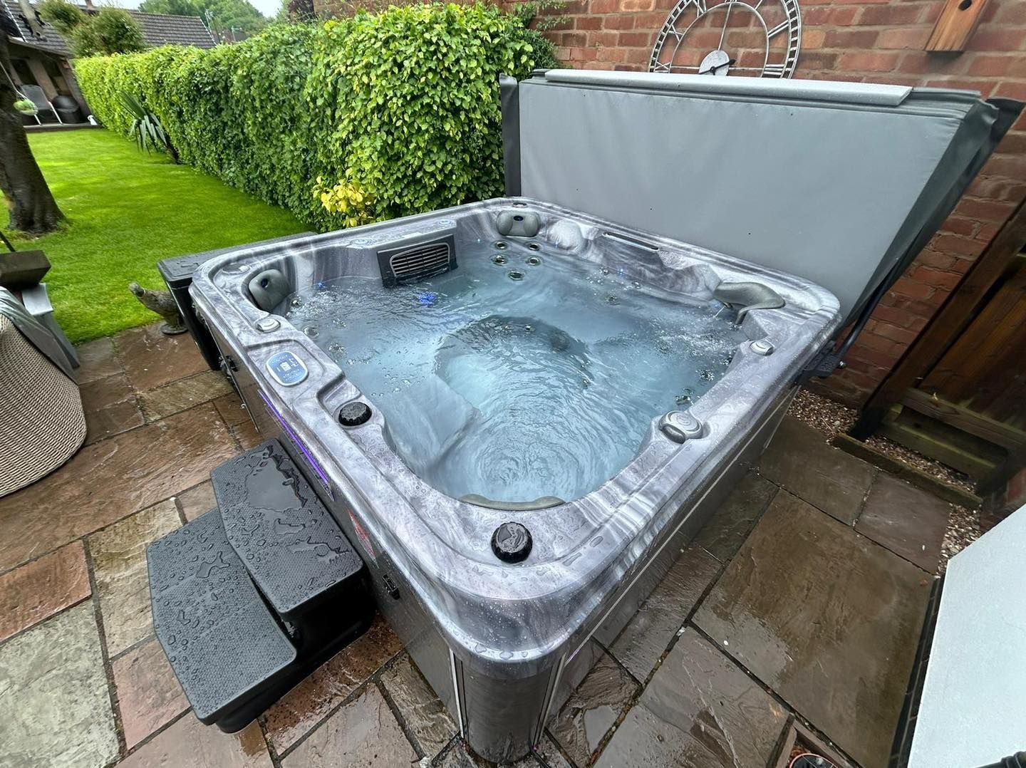 Hot tub filled with water on a stone patio, with steps leading up to it. Green hedge in background.
