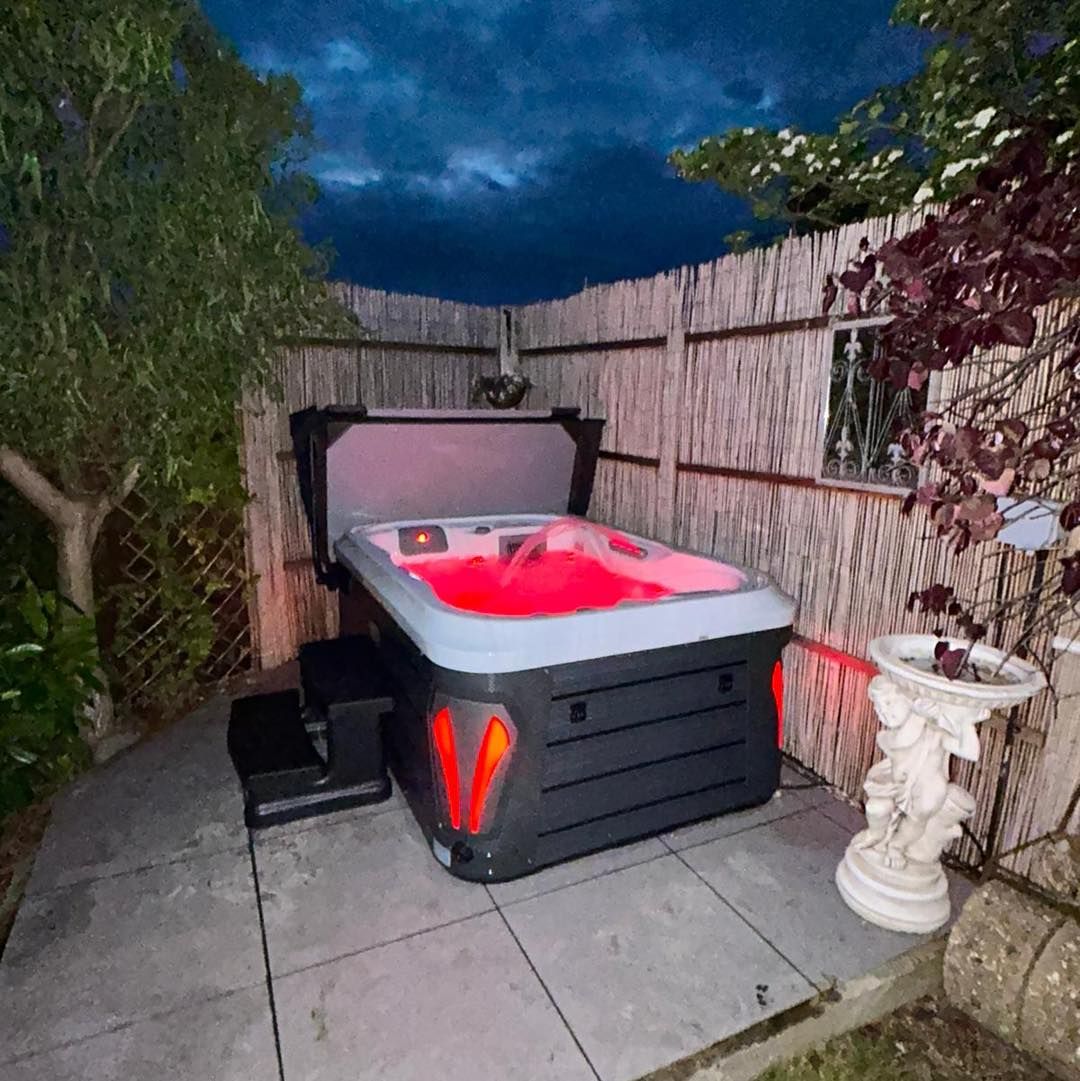 Hot tub with red interior lighting in an outdoor setting at dusk, surrounded by a fence and plants.