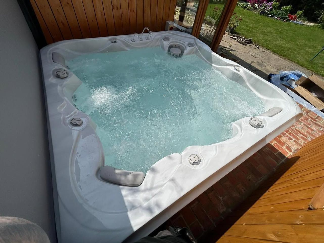 Square white hot tub with bubbling water in outdoor setting, framed by wooden structure.