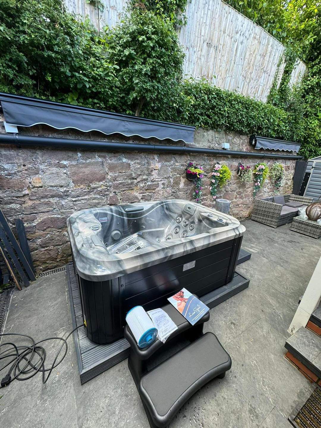 Hot tub on a patio. Black and gray tub with steps, stone wall, greenery, and awning.