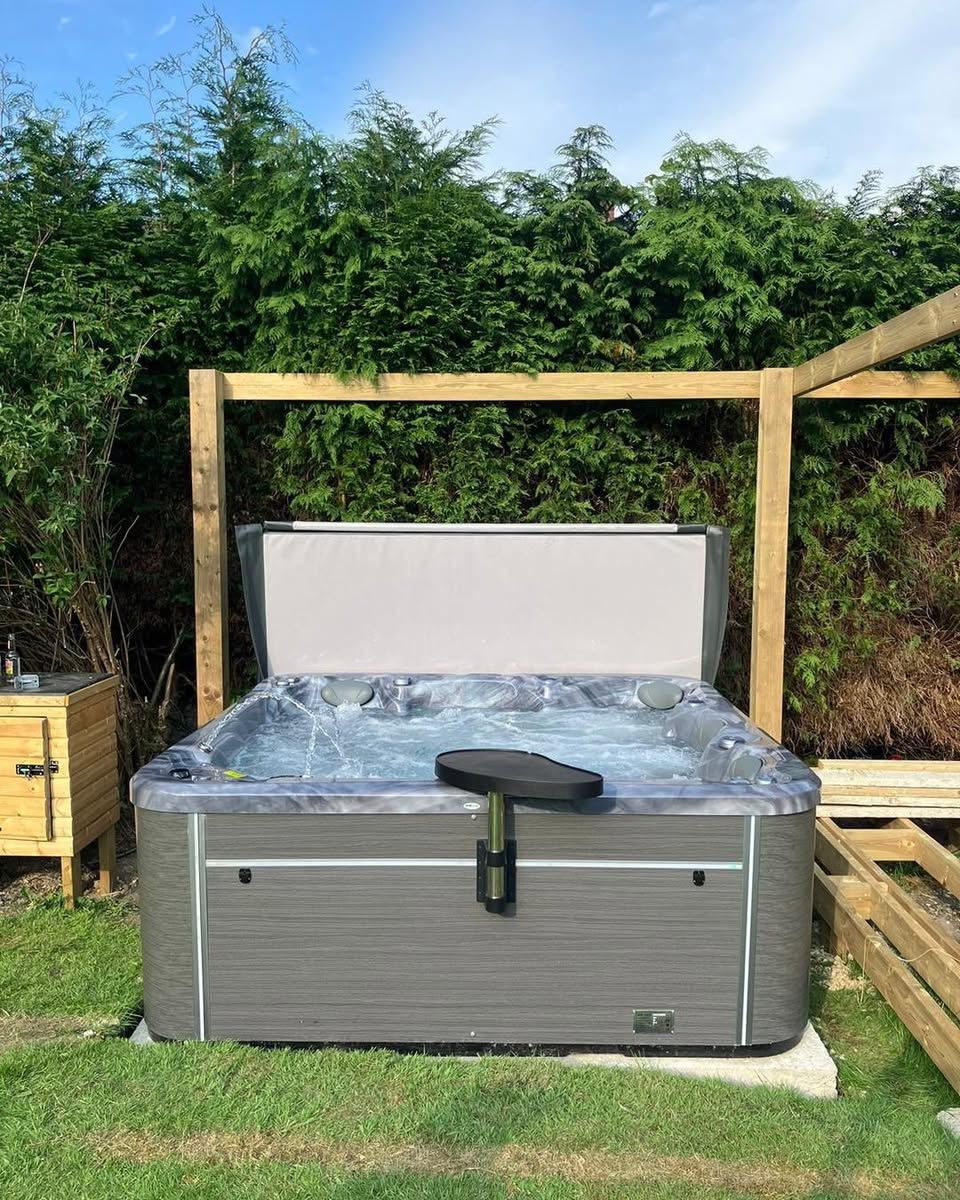 Hot tub with partially open cover, set on a lawn with a wooden frame and greenery in the background.