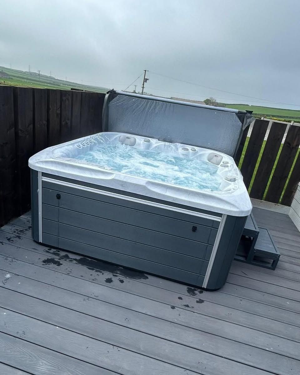 Hot tub on a wooden deck with cover open, bubbles visible, gray siding, and a cloudy sky.