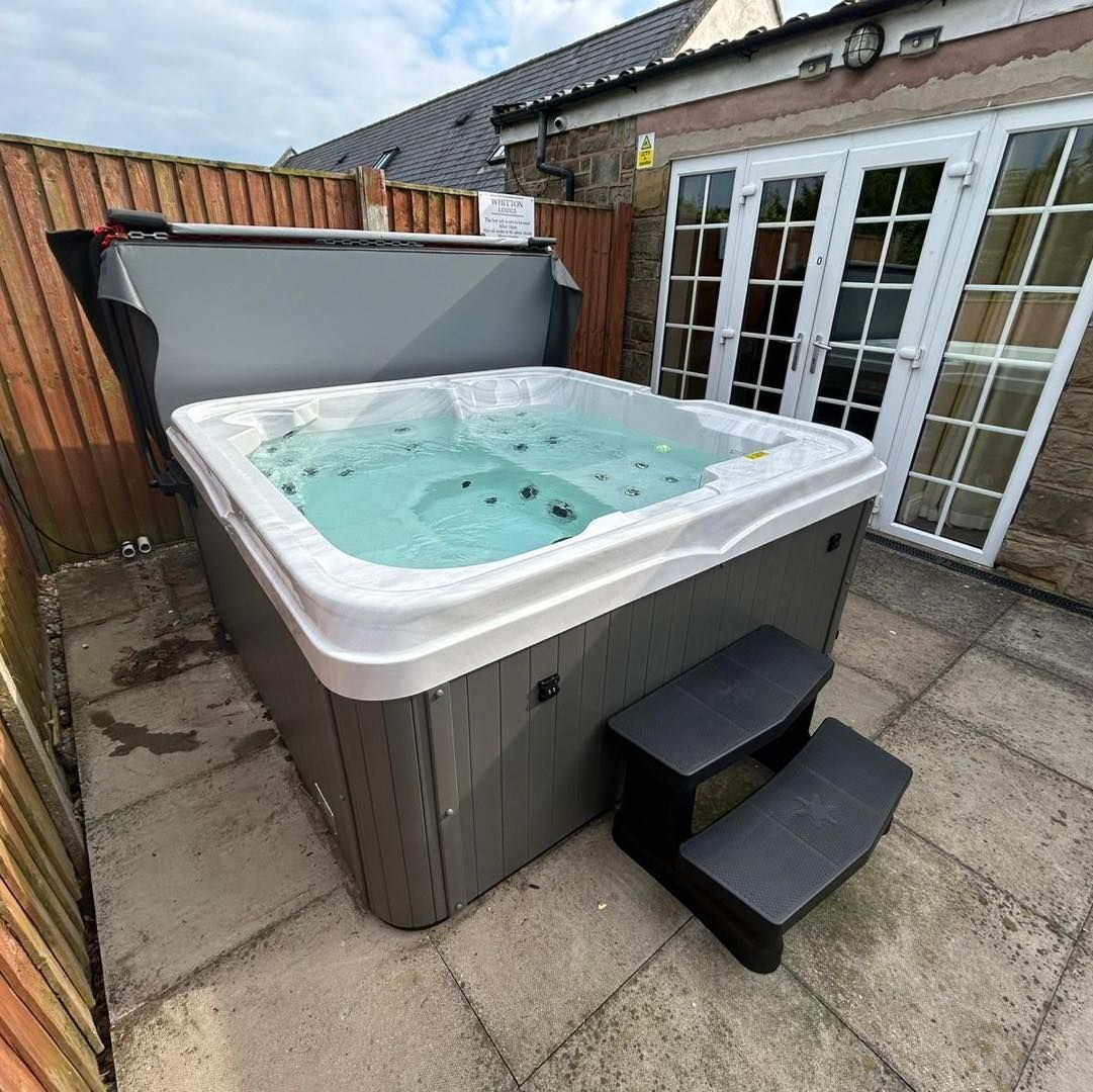 Hot tub with open cover, two-step entry, on patio. Grey tub, white trim, grey steps, near French doors and fence.