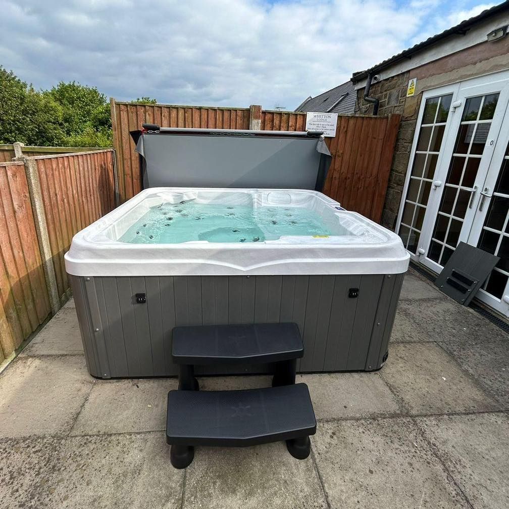 Hot tub with grey exterior, cover open, set on patio with steps, next to a fence.