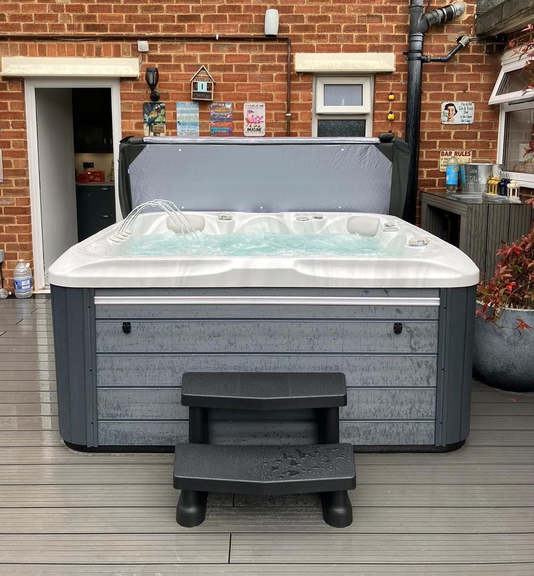 Hot tub with partially open cover, gray steps, on a wooden deck in front of a brick wall.
