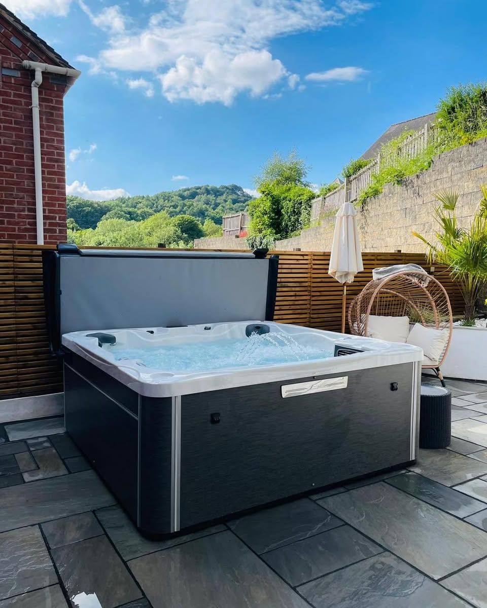 A hot tub with the cover partially open on a patio with a sunny, blue sky and a view of a hill.
