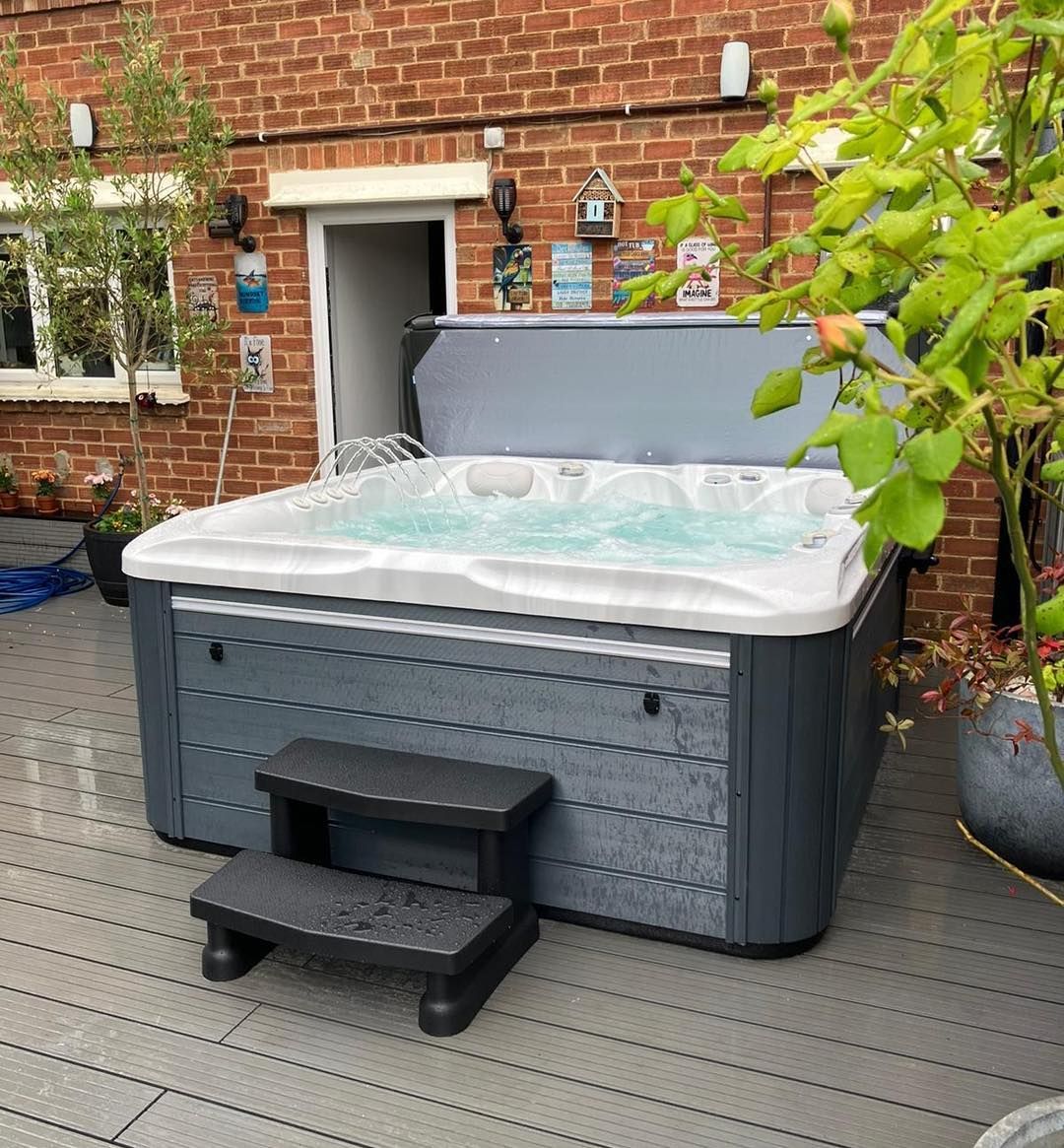 Hot tub on a grey deck against a brick wall, with open lid and steps.