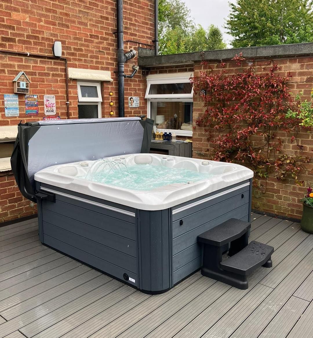 A dark gray hot tub with bubbling water on a gray deck, next to a brick wall with cover open and steps.