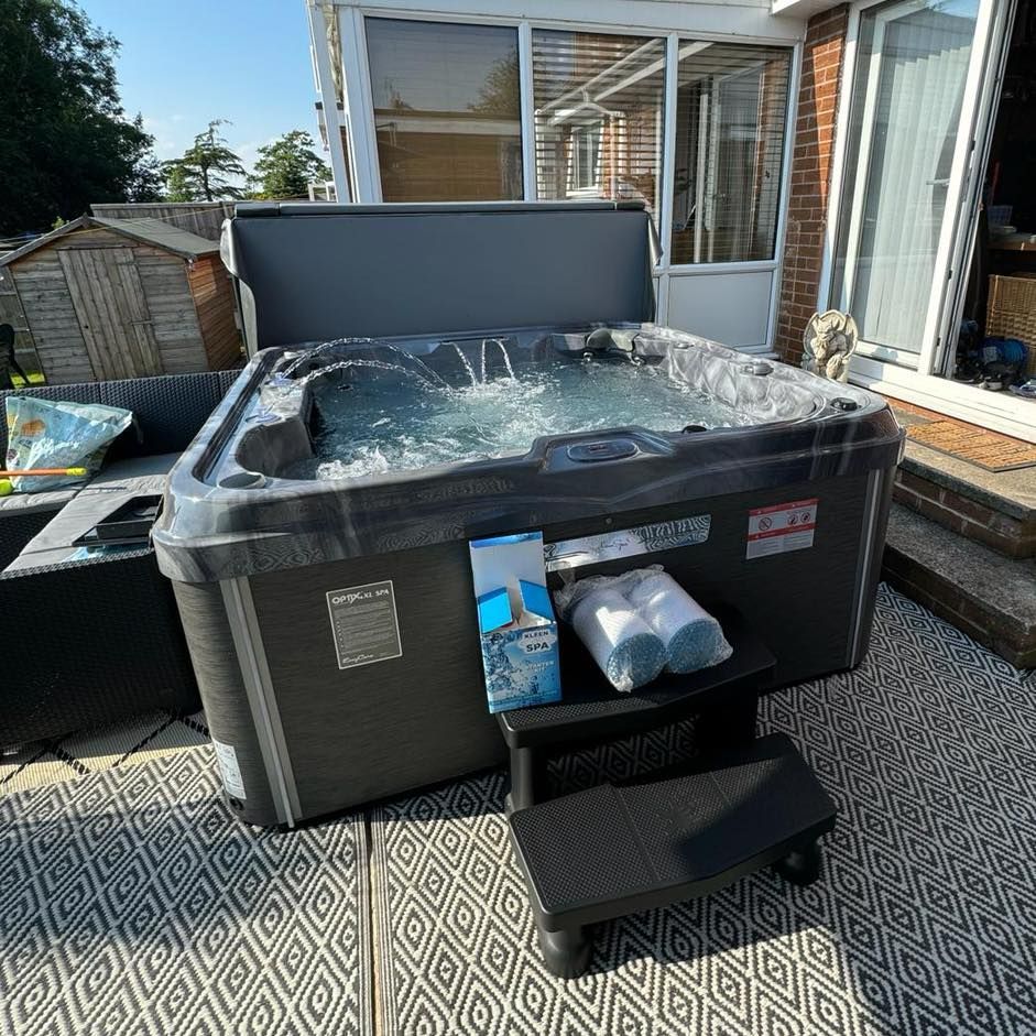 Hot tub with open cover, steps, and supplies on a patio with patterned rug.