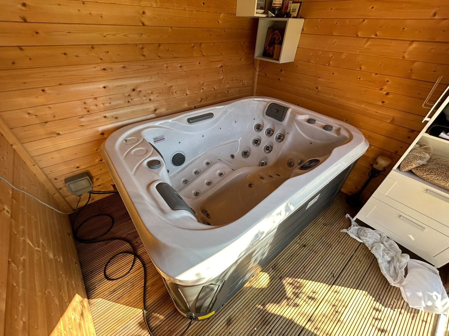 Hot tub inside a wooden-paneled room. The tub is light gray, with multiple jets. A cabinet and electrical outlet are visible.