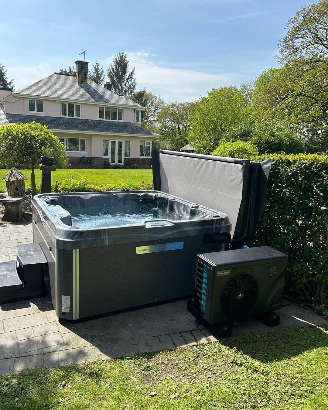 Hot tub on patio with open cover, next to pink house on sunny day.
