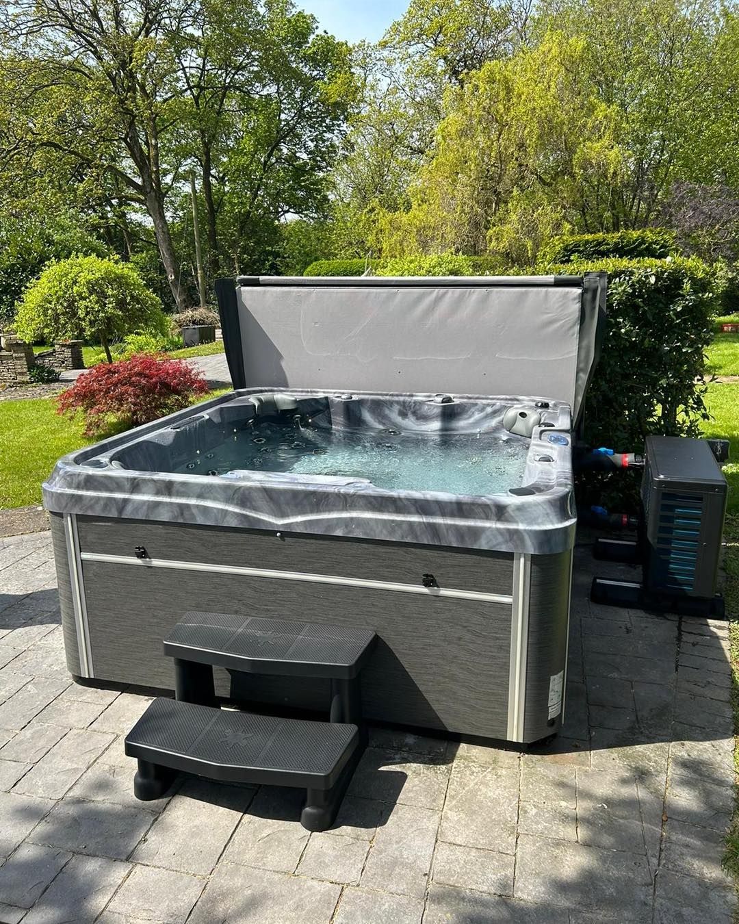 Hot tub with open cover, two-step entry, on stone patio. Lush green garden backdrop.