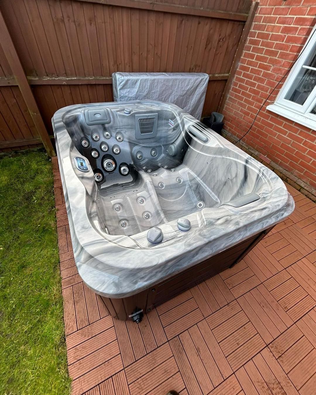 Hot tub on a wooden deck, near a wooden fence and brick wall. Gray and brown colors.