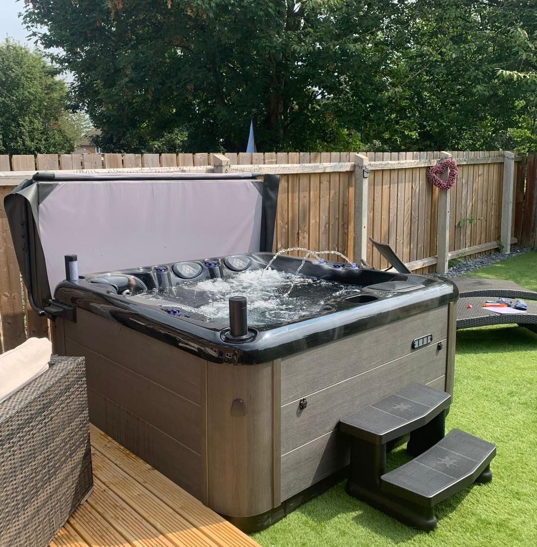 Hot tub with cover open, placed on wooden decking with steps, against a wooden fence and grass lawn.