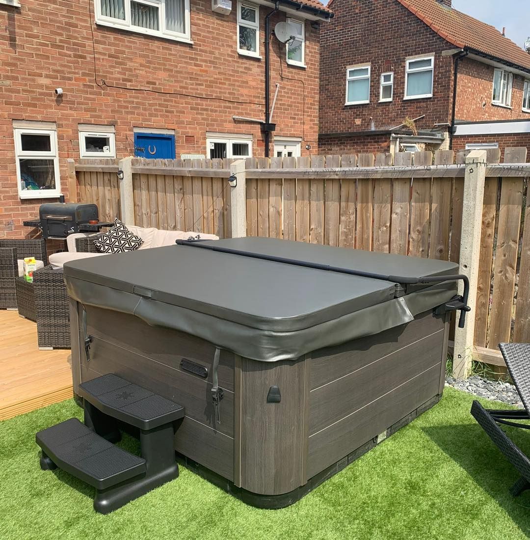 Hot tub with steps on a green lawn, near a wooden fence and houses.