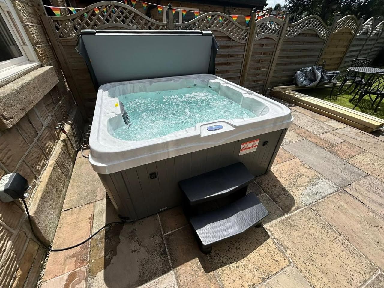 Hot tub with open cover on stone patio, steps in front. Fenced backyard with greenery.