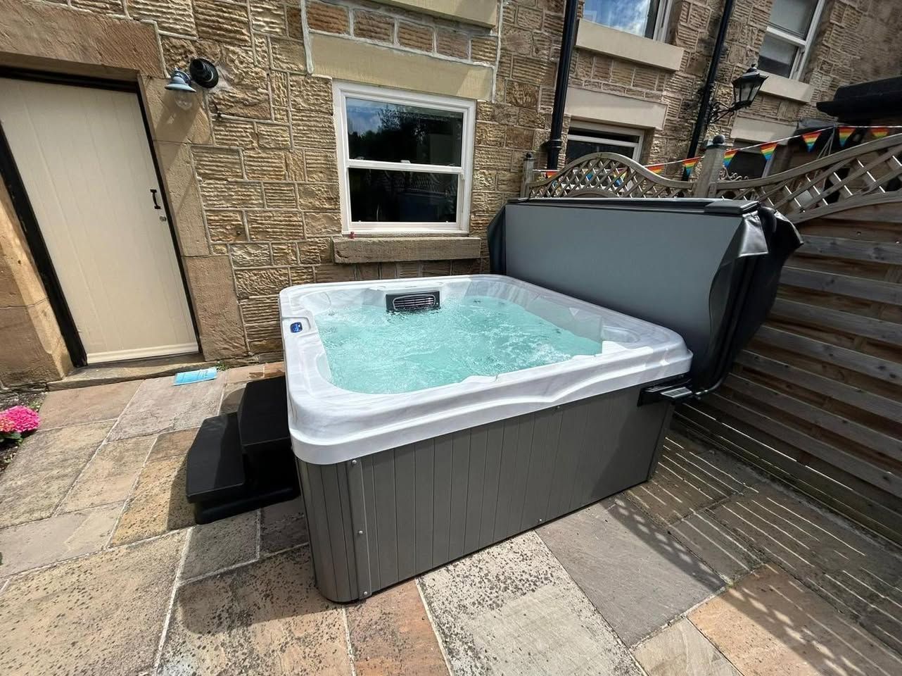 Hot tub with open cover on patio next to stone building with a door and window.