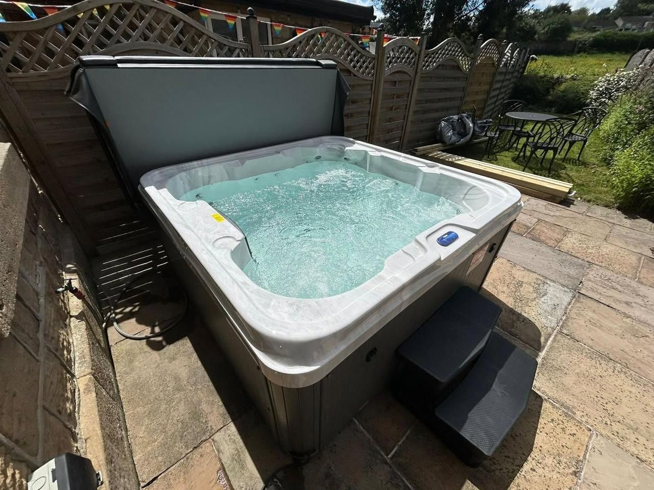Bubbling hot tub with open cover, on stone patio. Steps lead up to it, fenced yard in background.