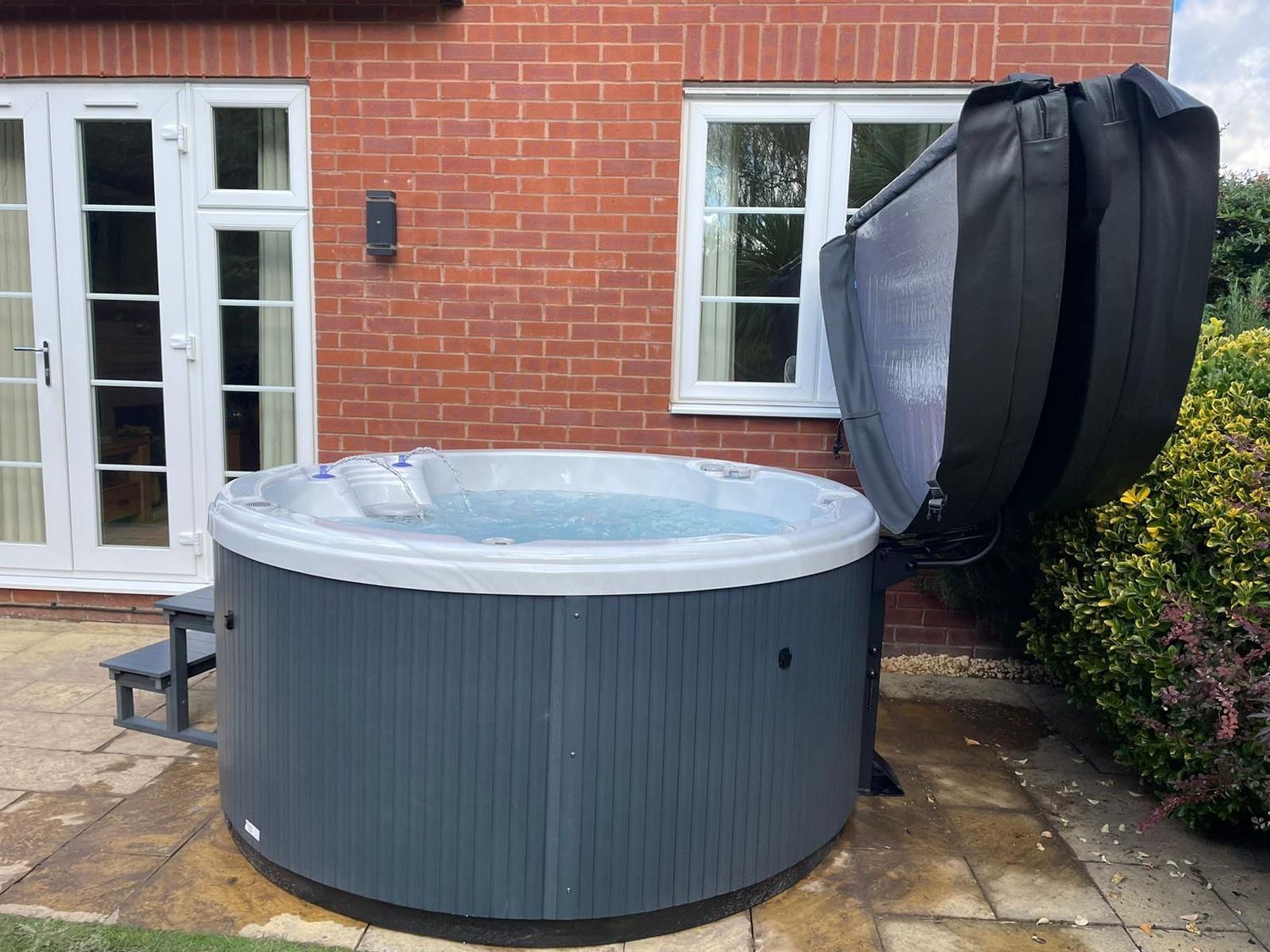 Round, grey hot tub with open black cover, on patio, brick wall background.