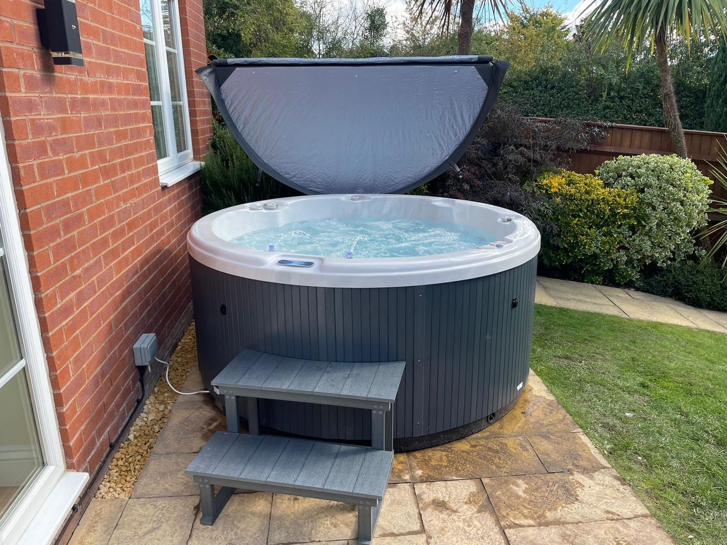 Hot tub outdoors with open gray lid, step, and brick building.