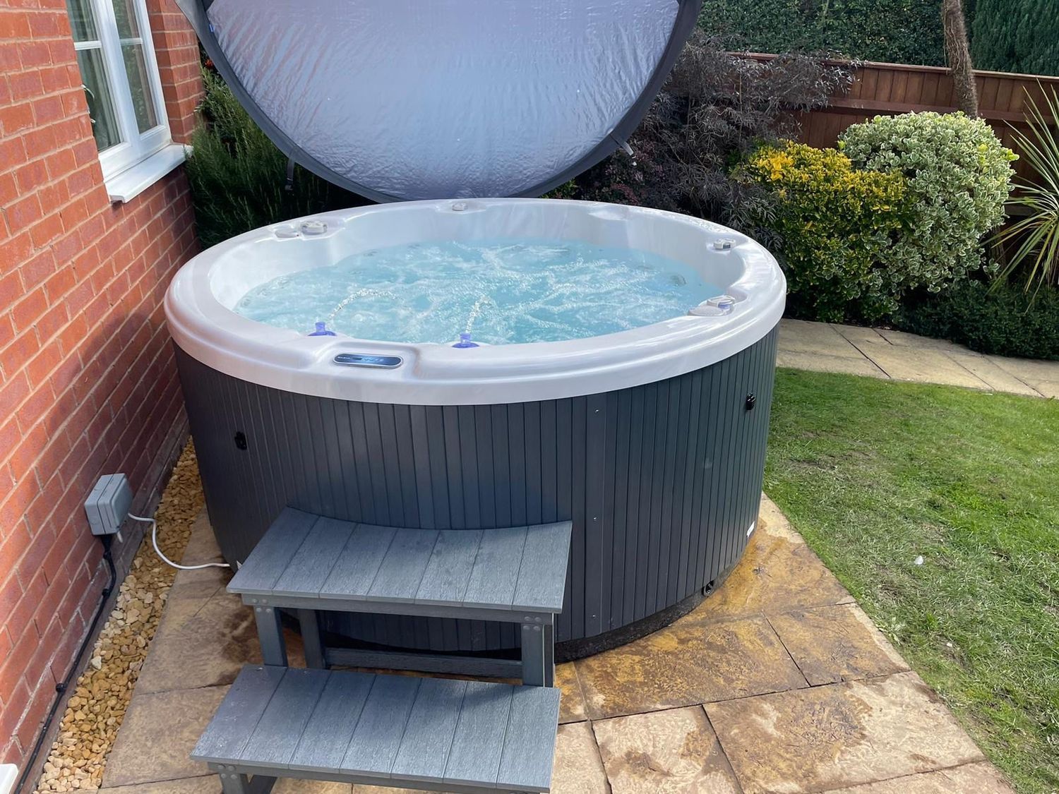 Hot tub with open cover in a backyard, beside a brick house. Grey steps provide access.