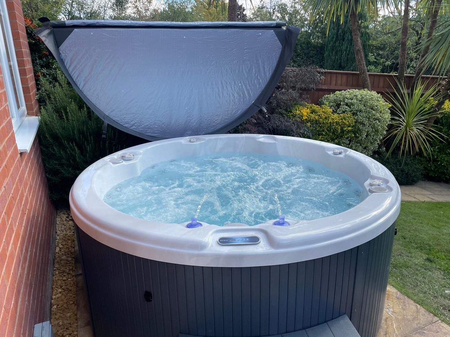 Round hot tub with water jets, cover partially open. Set outdoors near a brick wall and green foliage.