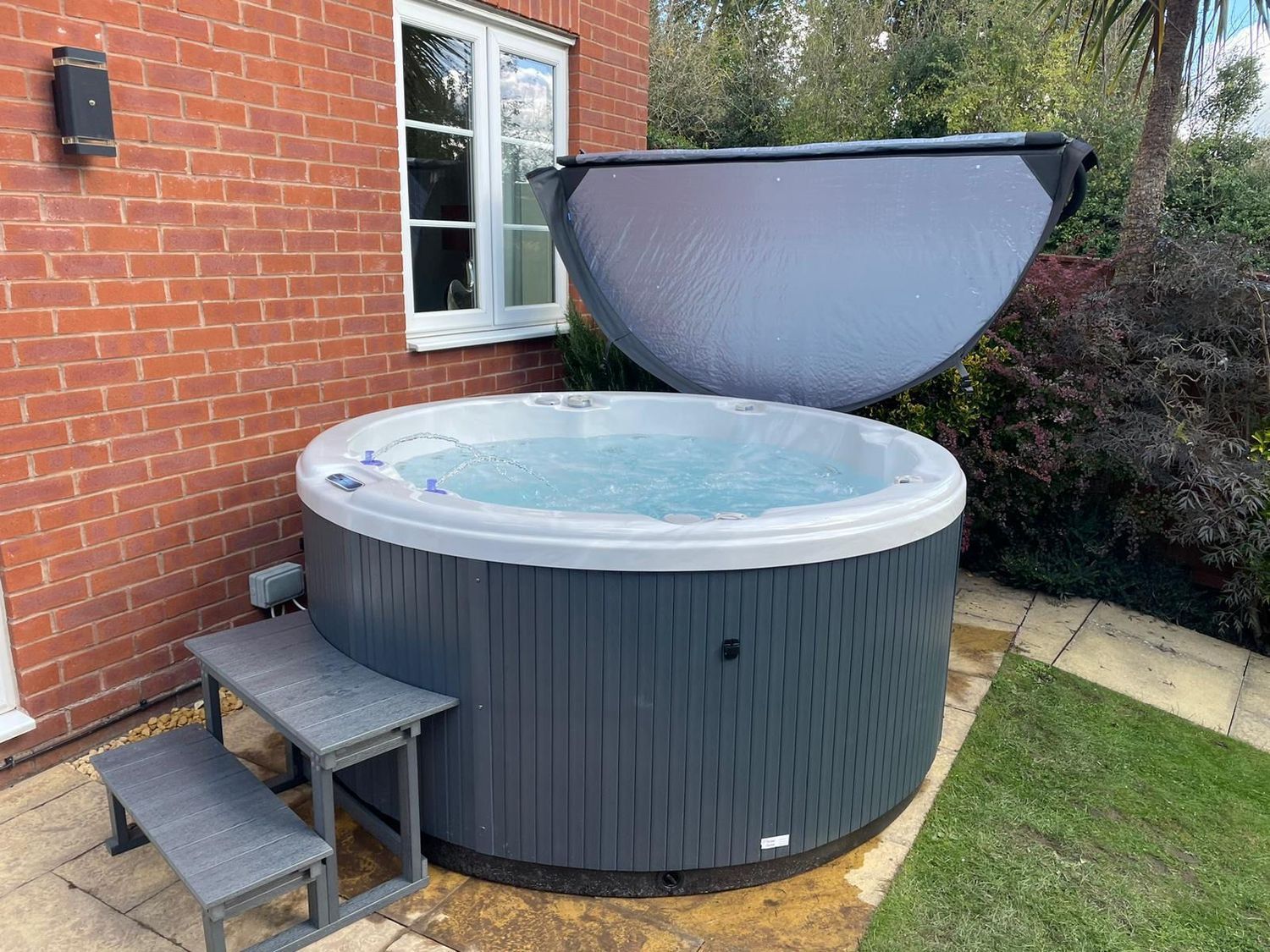 Round hot tub with raised steps, near a brick wall and green lawn, with open cover.