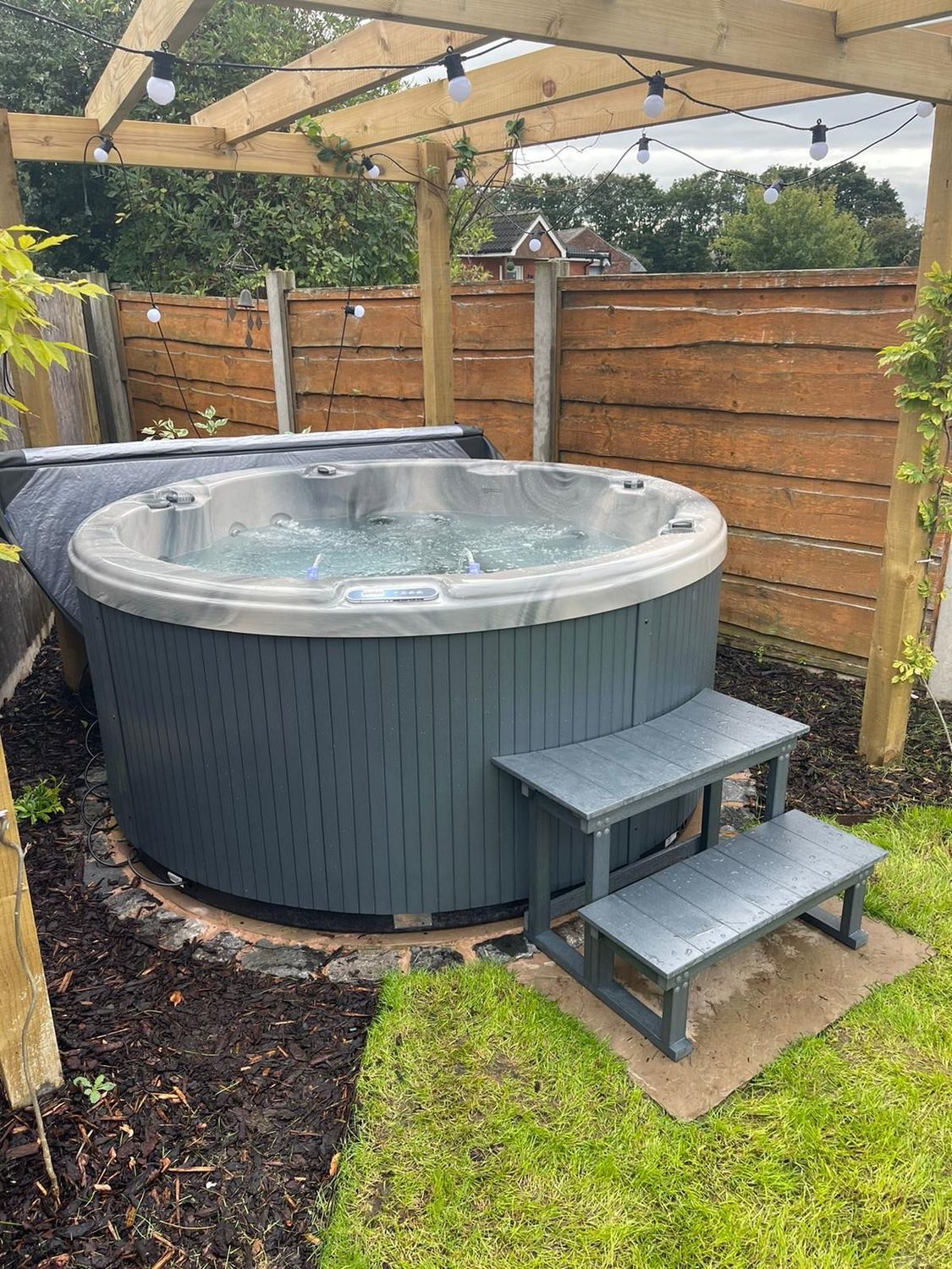 Round hot tub in a backyard with steps, under a wooden pergola and string lights.