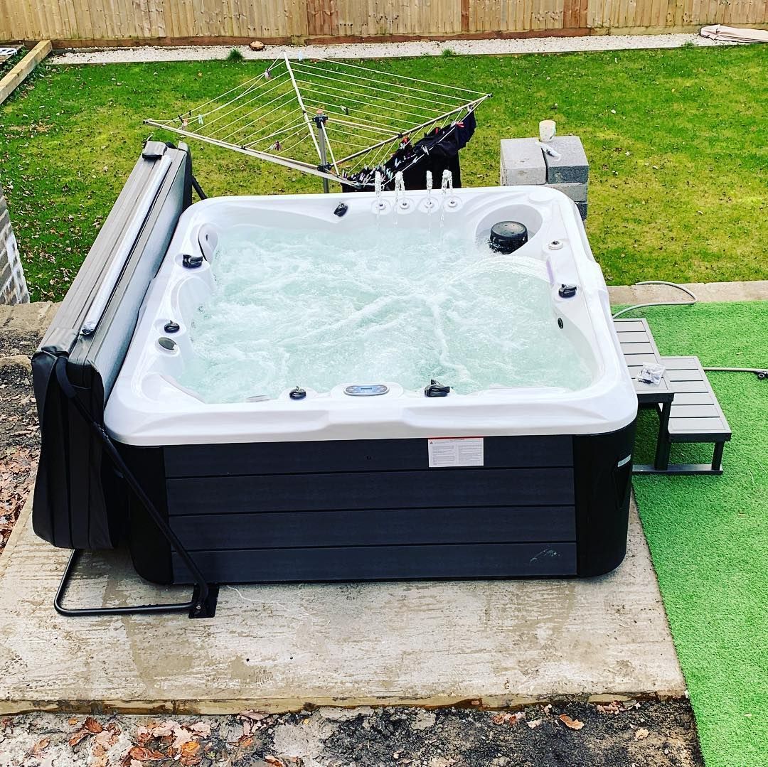 Hot tub with cover open on a concrete slab in a backyard. Bubbles in water. Steps and a clothes drying rack are nearby.