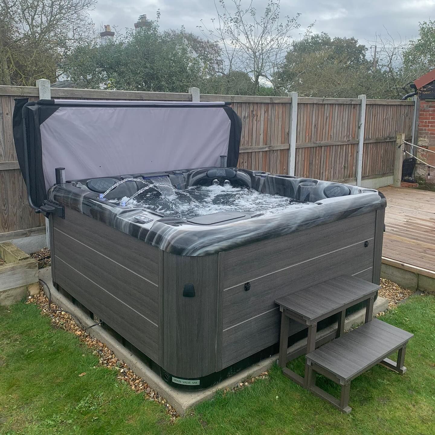 Hot tub with gray siding, open cover, bubbling water, and step stool on a patio.