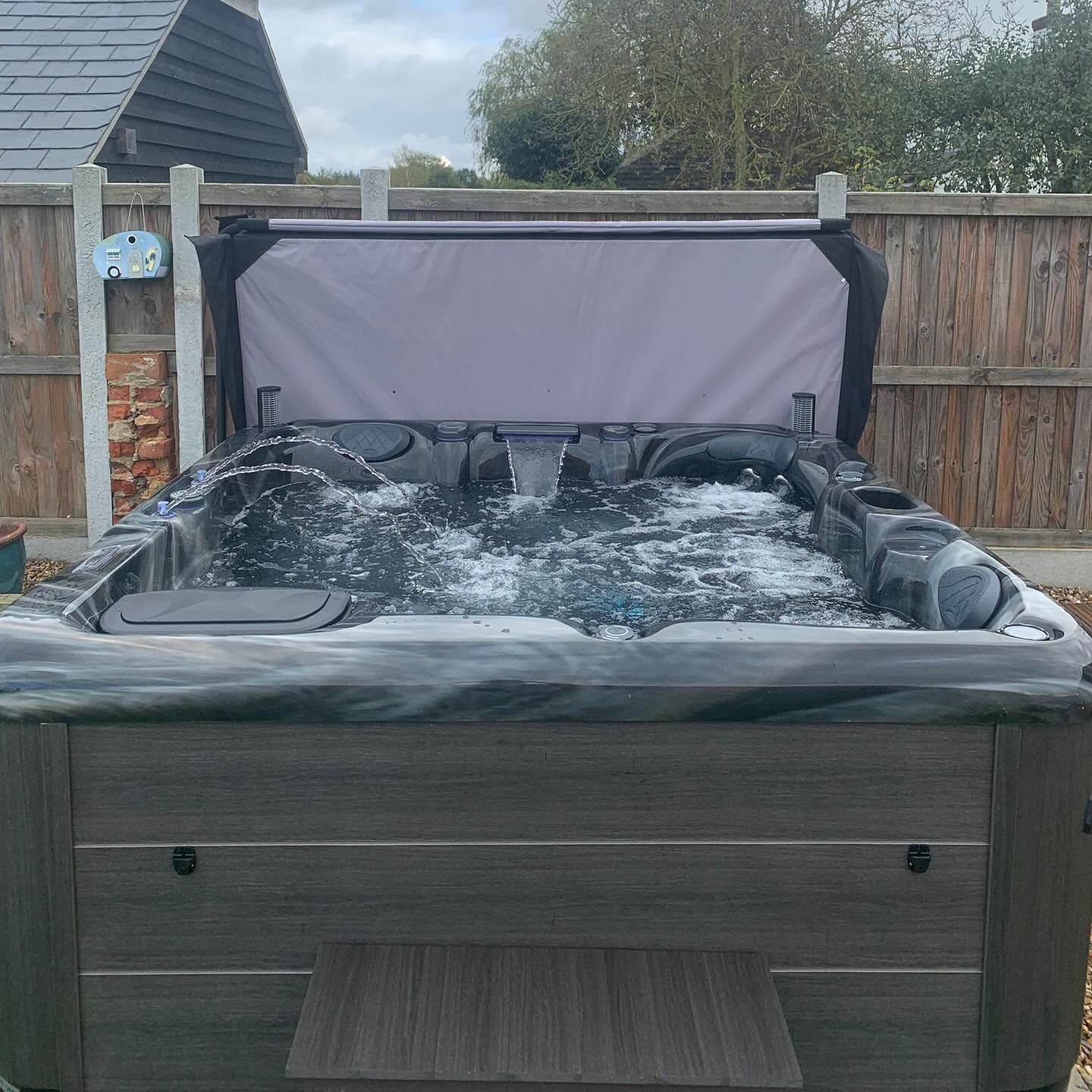 Hot tub with water jets active, cover partially open, outdoors next to wooden fence.