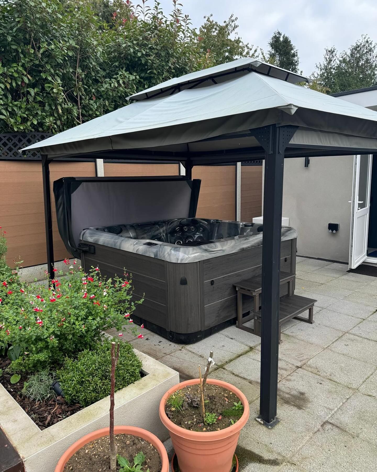 Hot tub under a gazebo on a patio. Brown tub, dark gray cover, plants nearby.
