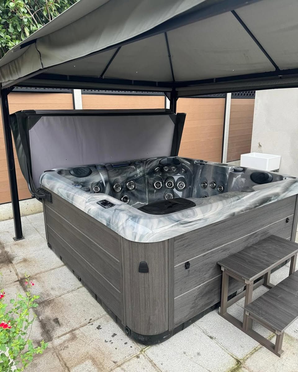 A hot tub with cover open, under a gray gazebo. There are steps, and a gray-brown facade.
