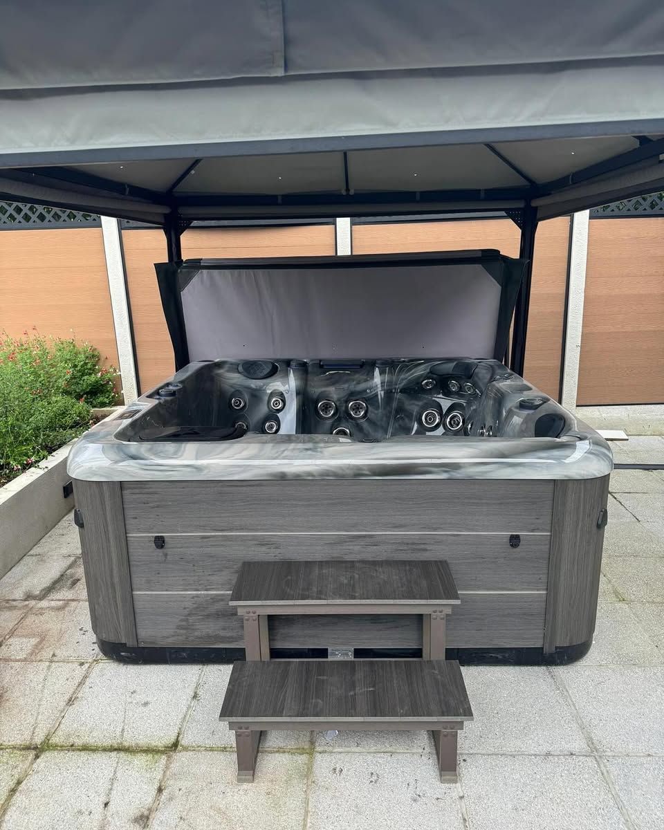 Hot tub under a gazebo, steps in front. Gray siding and cover. Outdoors.