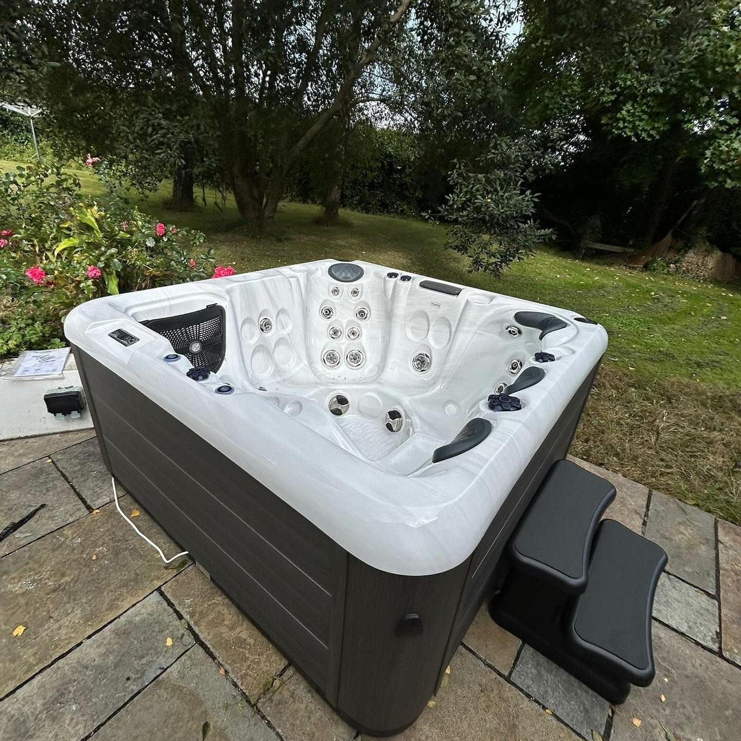 A dark gray hot tub with a white interior on a stone patio, steps to the right, green foliage in the background.