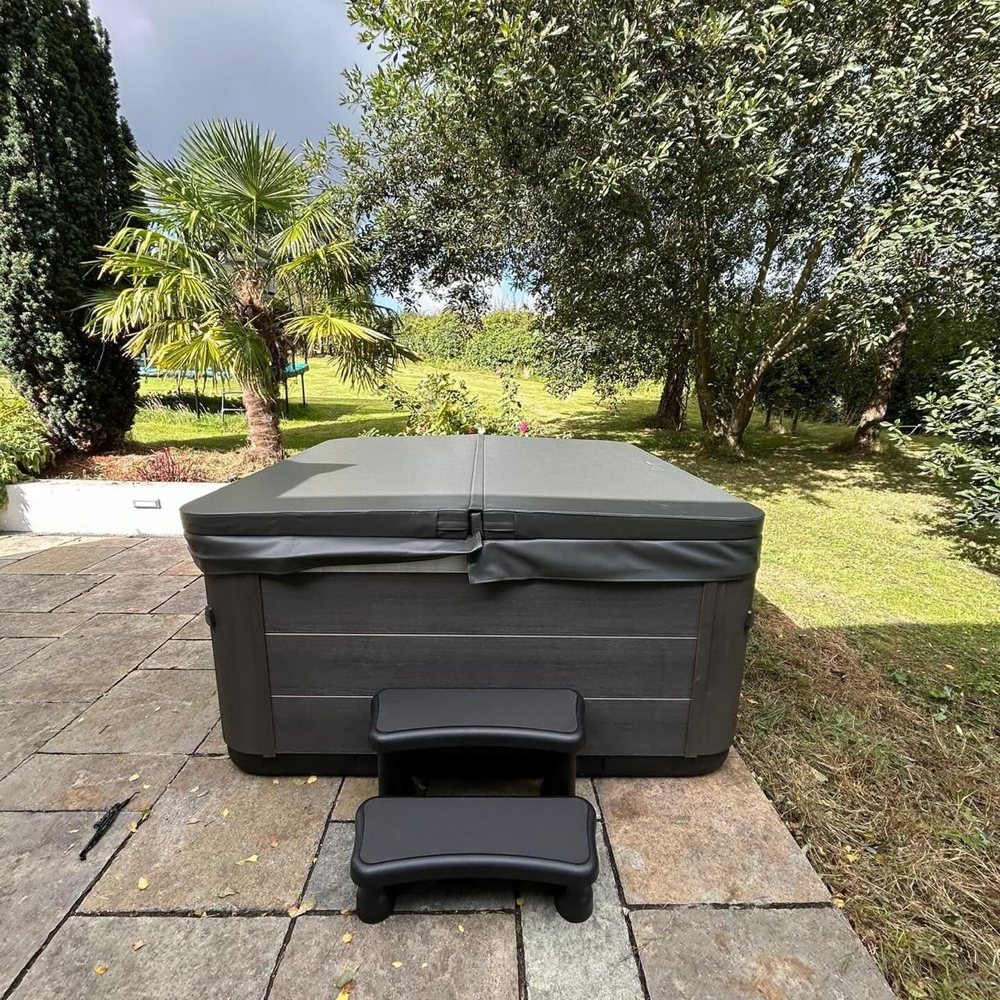 Hot tub with cover, black steps, on a stone patio, greenery in background.
