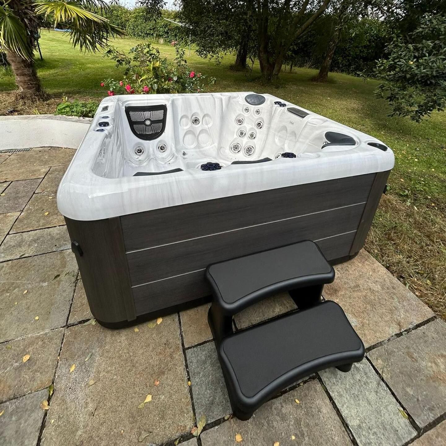 Hot tub with steps on a stone patio in a grassy yard. Gray-brown exterior, white interior.