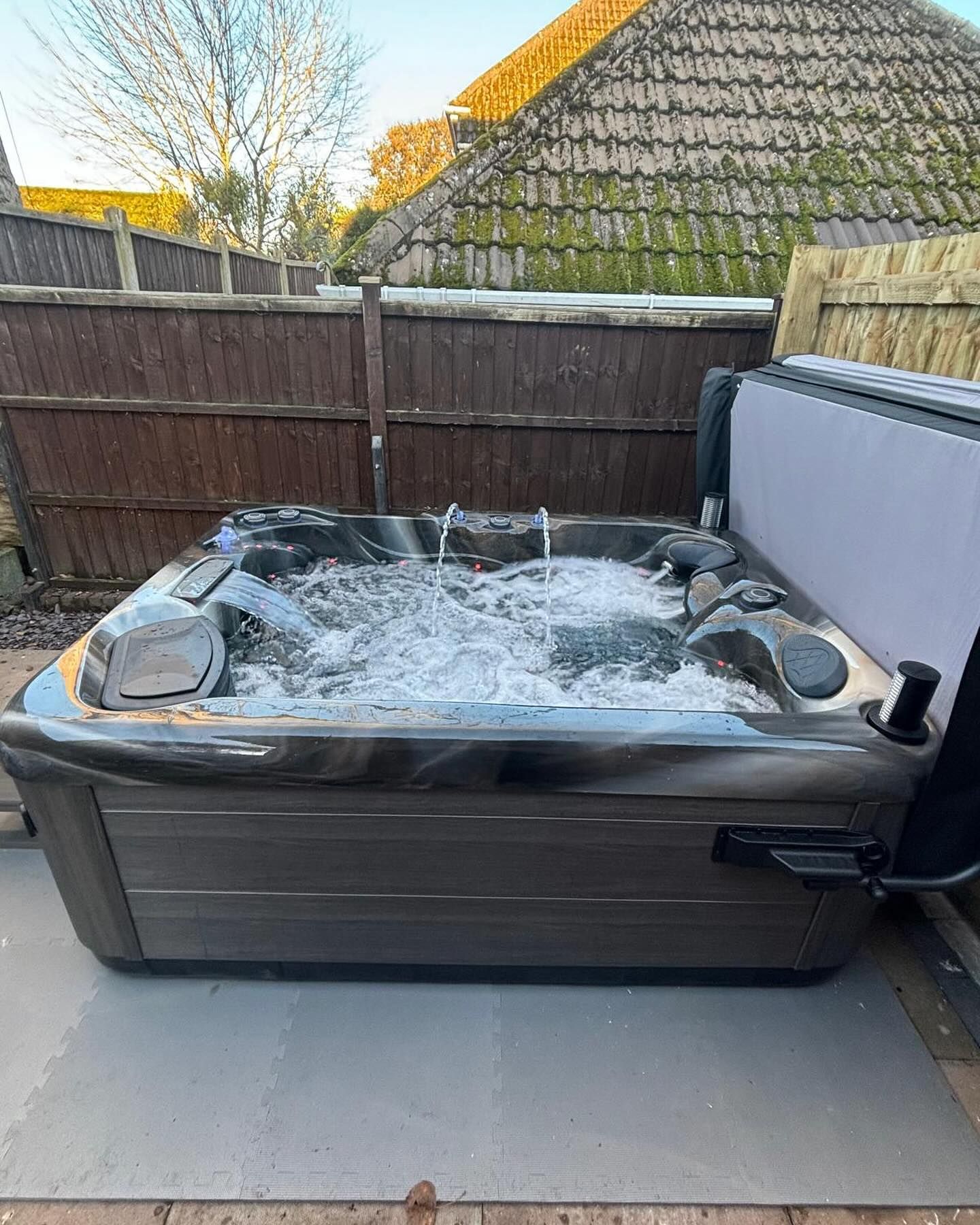 Hot tub with water jets, brown exterior, beside a fence in an outdoor setting.